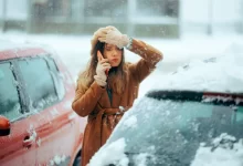 Žena si neví rady vedle auta v zimě a telefonuje.