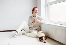Žena sedí u radiátoru se svým psem a pije čaj z hrníčku.