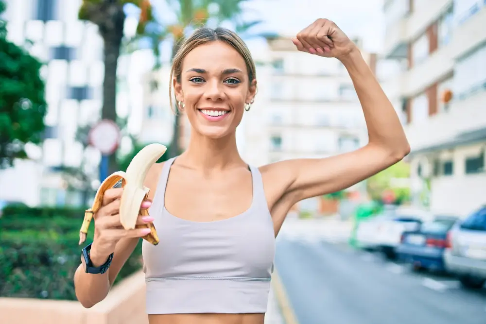 shutterstock_1932454319 - SportDeník Mladá žena drží v ruce banán, zatíná biceps a směje se.