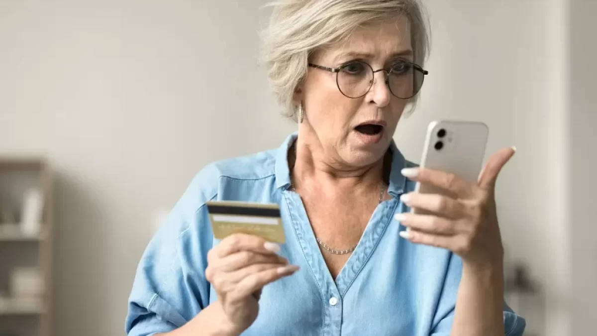 Starší žena se zděšeně dívá na svůj mobilní telefon a kreditní kartu