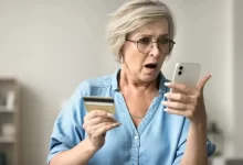 Starší žena se zděšeně dívá na svůj mobilní telefon a kreditní kartu