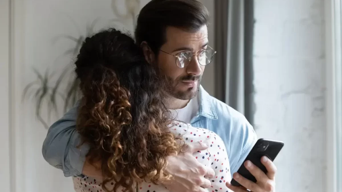 Muž drží ženu kolem ramen a přitom se dívá do mobilního telefonu