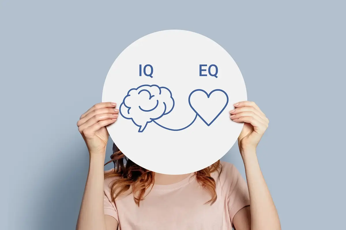 Rébus 4 - SportDeník Mladá žena drží před obličejem tabulku se zkratkami IQ a EQ