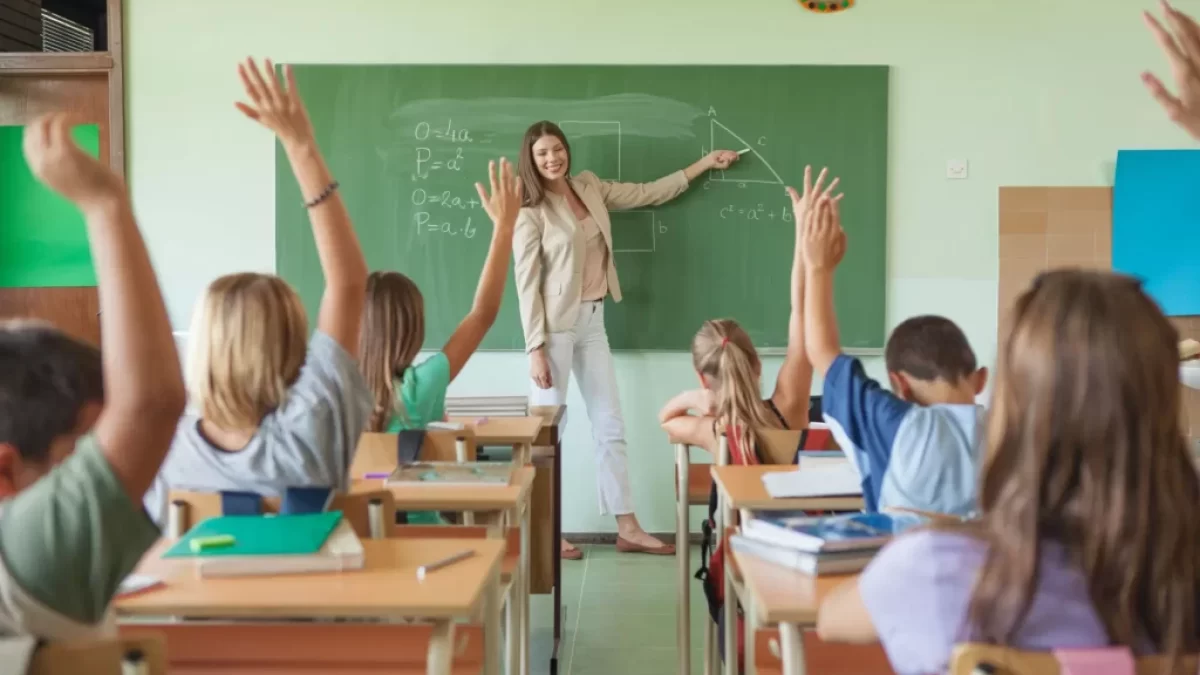 Třída s hlásícími se žáky a paní učitelkou před tabulí