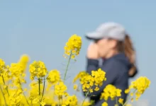 Mladá žena stojí v pozadí za rozkvetlým kečem a drží si u nosu kapesník