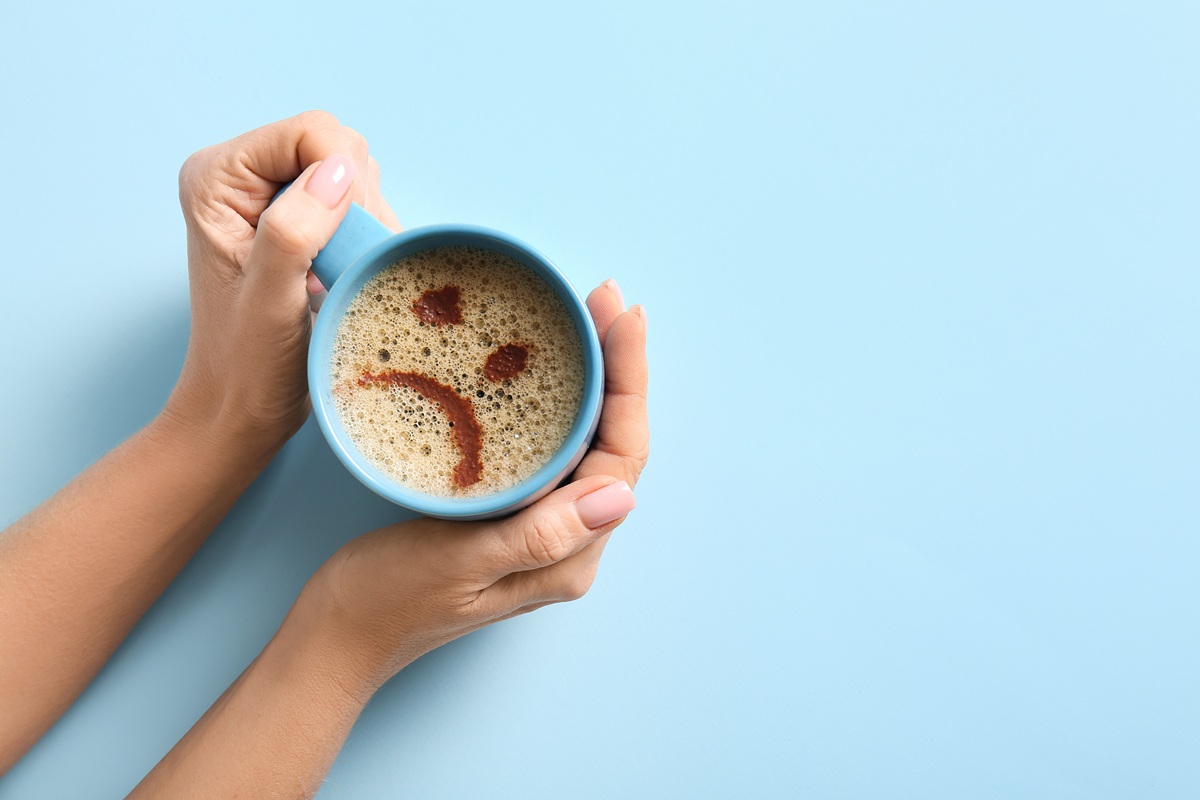 HandsHoldingCupOfCoffeeWithDrawnSadFaceTop - SportDeník Hrnek s kávou a zamračeným smajlíkem
