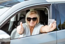 Starší žena řidička ukazuje palec nahoru z auta, v ruce drží řidičský průkaz