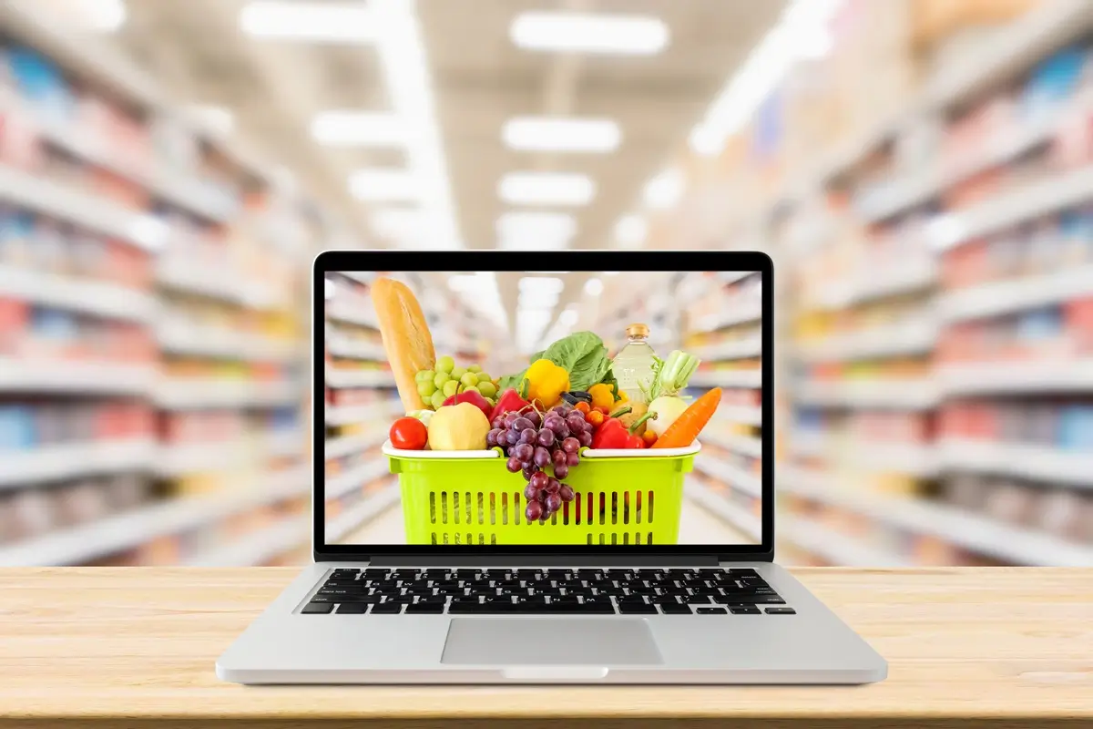 Pohled do regálů supermarketu, v popředí je košík se zeleninou a jídlem na obrazovce noteboooku.