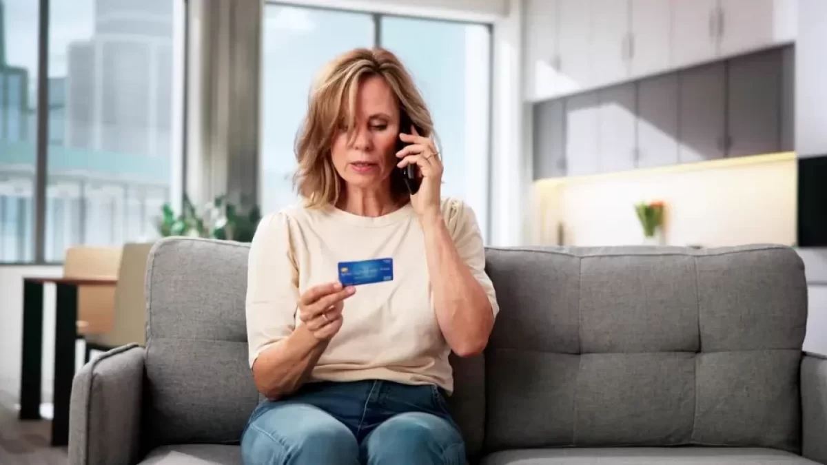 Žena středního věku sedící na sedačce s mobilním telefonem u ucha a platební kartou v ruce