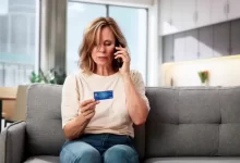 Žena středního věku sedící na sedačce s mobilním telefonem u ucha a platební kartou v ruce