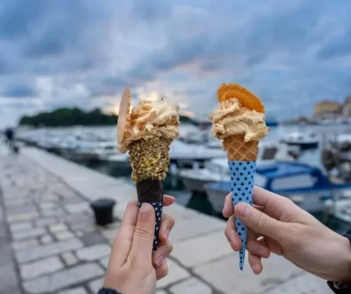 Promenáda u moře v Chorvatsku a před ní ve vzduchu dvě ruce s kornoutem zmrzliny