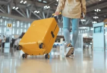 Mladý muž v odbavovací hale na letišti táhne za sebou velký žlutý kufr na kolečkách