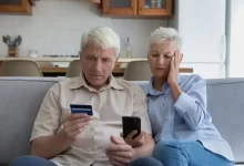 Starší manželský pár hledí nevěřícně na svou platební kartu a do mobilního telefonu