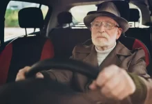 Starší muž v klobouku a brýlích soustředěně drží volant v autě.