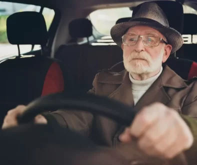 Starší muž v klobouku a brýlích soustředěně drží volant v autě.