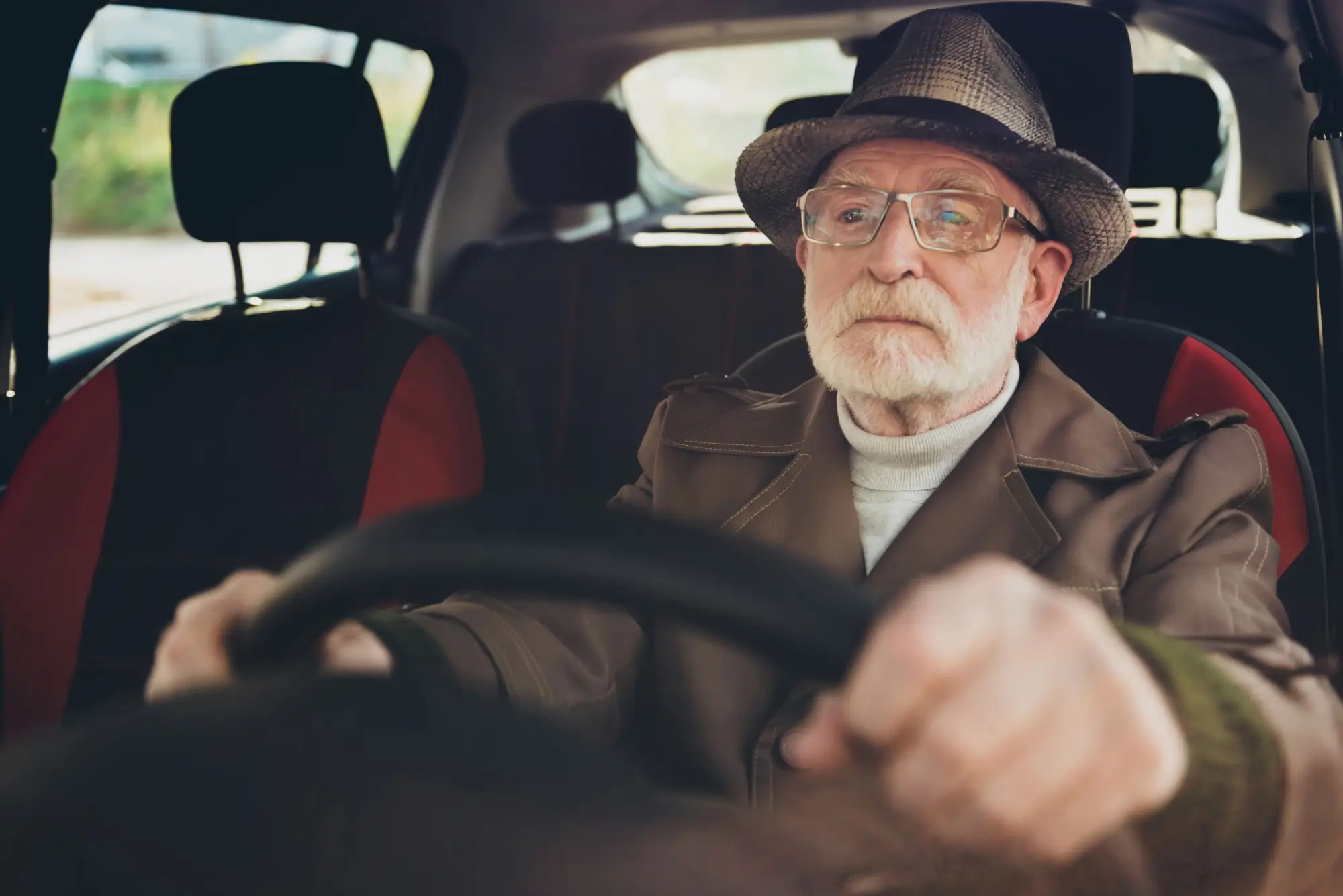 Starší muž v klobouku a brýlích soustředěně drží volant v autě.