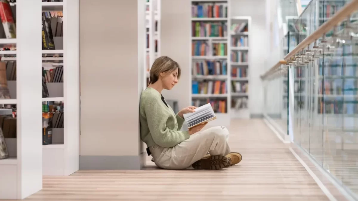 Studentka sedí v knihovně na zemi mezi regály a čte si