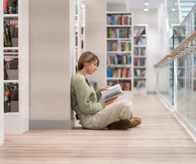 Studentka sedí v knihovně na zemi mezi regály a čte si
