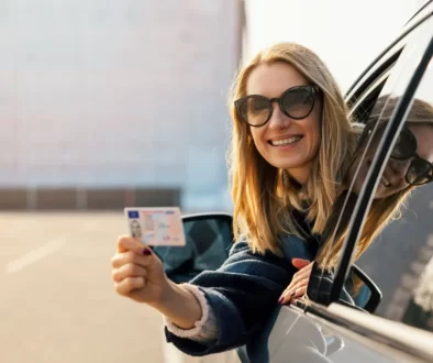 Žena v autě se usmívá a ukazuje řidičský průkaz.