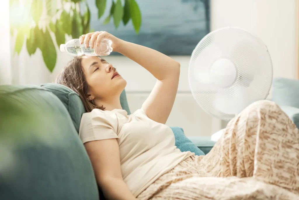 Shutterstock_2529706947 - SportDeník Žena doma chladící hlavu lahví s vodou při letním vedru, vedle ní běží ventilátor.
