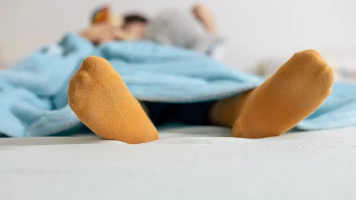 Osoba ležící pod modrou přikrývkou s oranžovými ponožkami.