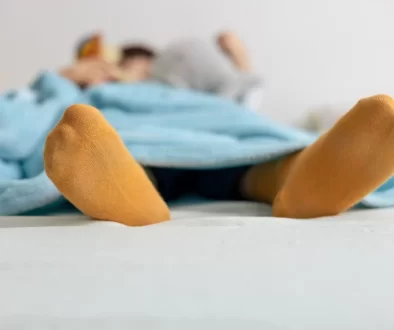 Osoba ležící pod modrou přikrývkou s oranžovými ponožkami.