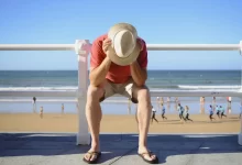 Turista sedící na promenádě se sklopenou hlavou u pláže, v pozadí lidé u moře.