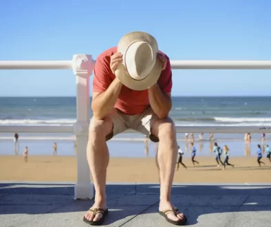 Turista sedící na promenádě se sklopenou hlavou u pláže, v pozadí lidé u moře.