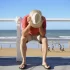Turista sedící na promenádě se sklopenou hlavou u pláže, v pozadí lidé u moře.