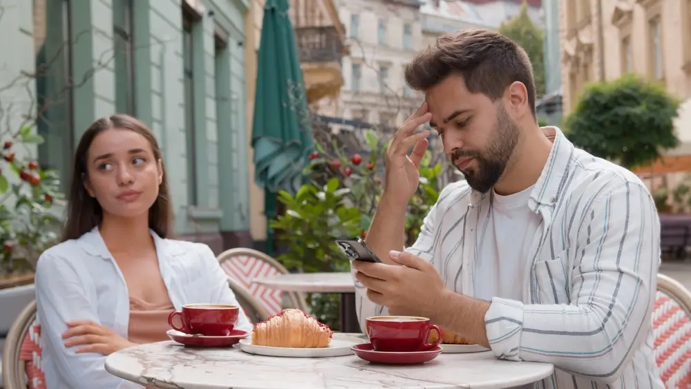 shutterstock_2434921813 - SportDeník Pár sedí u stolu, muž má v ruce telefon