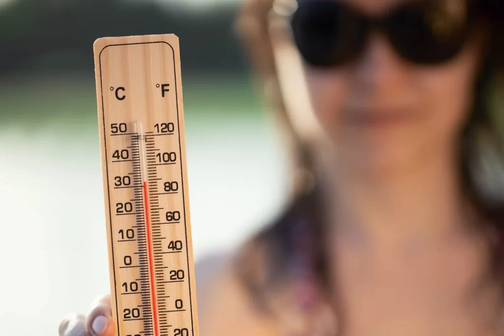 - SportDeník Detail dřevěného teploměru ukazujícího teplotu nad 30 °C, žena v pozadí s tmavými brýlemi během slunečného dne.