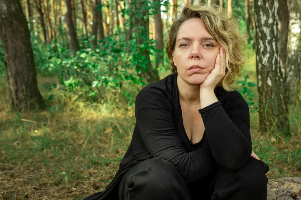 Žena v černém oblečení sedící zamyšleně v lese, opřená o ruku, s vážným výrazem.