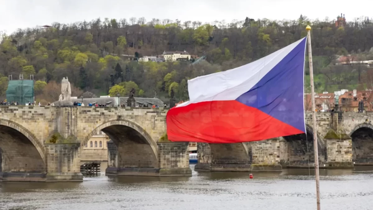 Česká vlajka vlající na stožáru s výhledem na Karlův most a panorama historického centra Prahy při západu slunce.