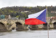 Česká vlajka vlající na stožáru s výhledem na Karlův most a panorama historického centra Prahy při západu slunce.