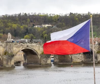 Česká vlajka vlající na stožáru s výhledem na Karlův most a panorama historického centra Prahy při západu slunce.