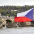 Česká vlajka vlající na stožáru s výhledem na Karlův most a panorama historického centra Prahy při západu slunce.