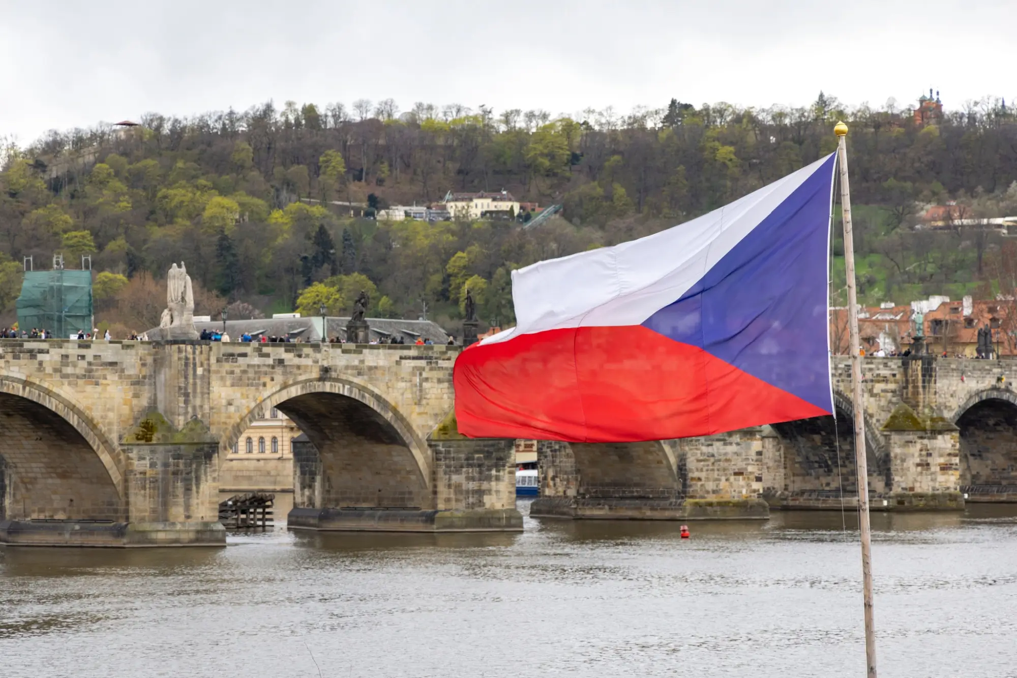Česká vlajka vlající na stožáru s výhledem na Karlův most a panorama historického centra Prahy při západu slunce.