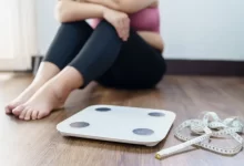 Žena sedí na podlaze vedle váhy a svinutého metru, v zamyšlené nebo nejisté náladě.