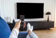 Muž na gauči drží ovladač namířený na televizi