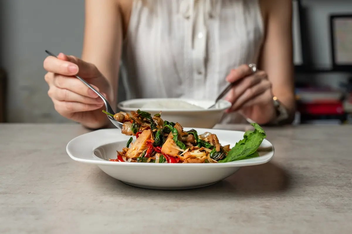 Mladá žena sedí za stolem, v ruce drží příbor a chystá se jíst krásně barevné veganské jídlo