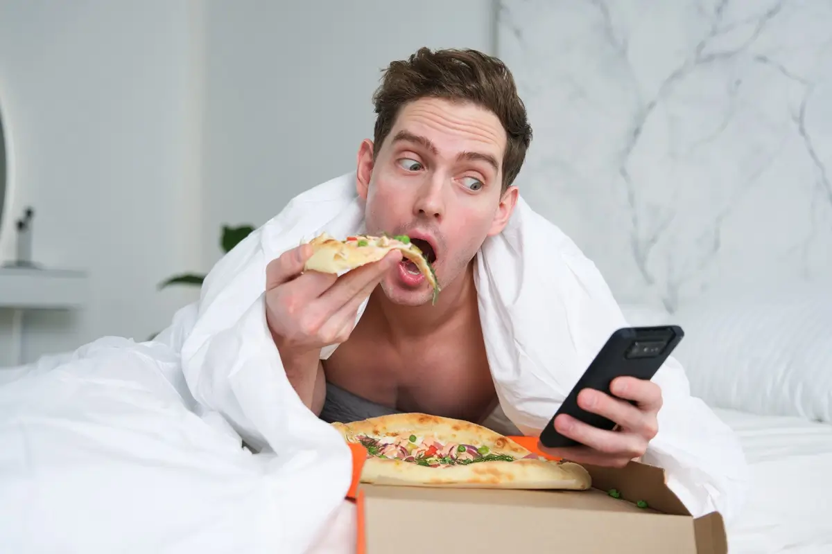 Mladý atraktivní muž se cpe v posteli kouskem pizzy, další má v krabici před sebou. Dívá se u toho na telefon. 