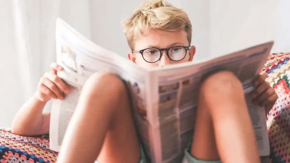 Asi desetiletý blonďatý chlapec sedí na sedačce a půl obličeje mu zakrývá časopis