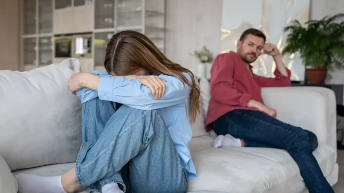 Mladý pár sedící zády k sobě na sedačce, žena si zakrývá obličej, muž se dívá na ni