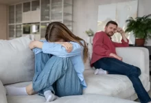 Mladý pár sedící zády k sobě na sedačce, žena si zakrývá obličej, muž se dívá na ni