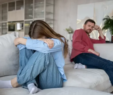 Mladý pár sedící zády k sobě na sedačce, žena si zakrývá obličej, muž se dívá na ni