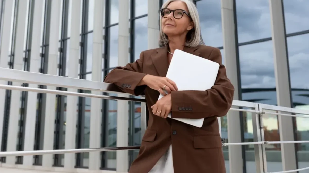 Žena 60+ v hnědém saku a papíry v ruce stojí před prosklenou budovou