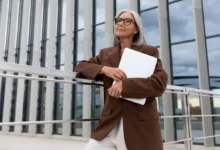 Žena 60+ v hnědém saku a papíry v ruce stojí před prosklenou budovou