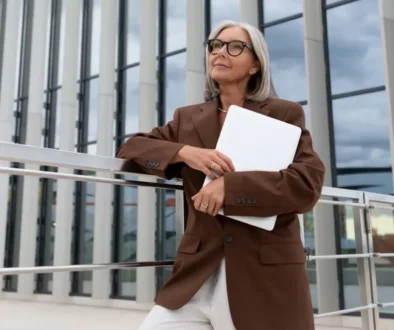 Žena 60+ v hnědém saku a papíry v ruce stojí před prosklenou budovou