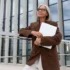 Žena 60+ v hnědém saku a papíry v ruce stojí před prosklenou budovou