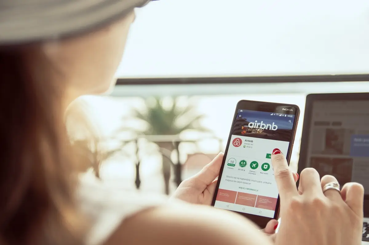 Mladá žena v klobouku si přes telefon objednává ubytování na platformě Airbnb